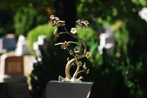 Abstract gold sculpture on a black pedestal outdoors with blurred greenery in the background
