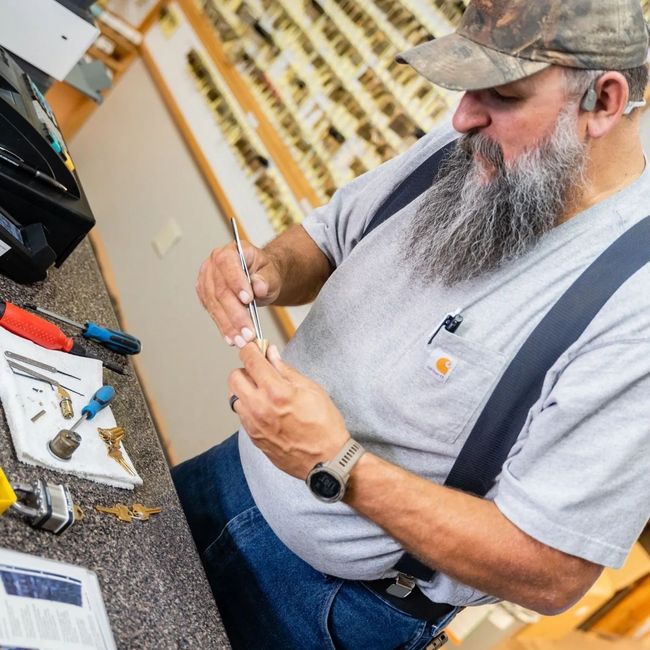 A bearded locksmith in a cap and gray shirt works on a lock component at a counter in a shop filled with keys.