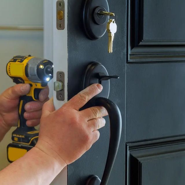 A person uses a yellow cordless drill to install a black door handle and deadbolt on a dark-painted door.
