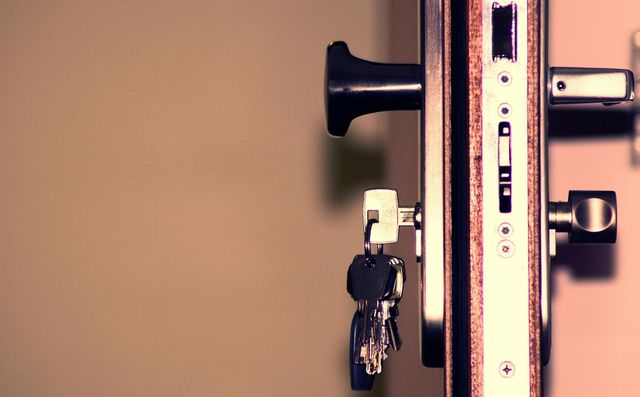 A metal door lock mechanism with a set of keys hanging in the keyhole, shown in profile against a plain, light background.