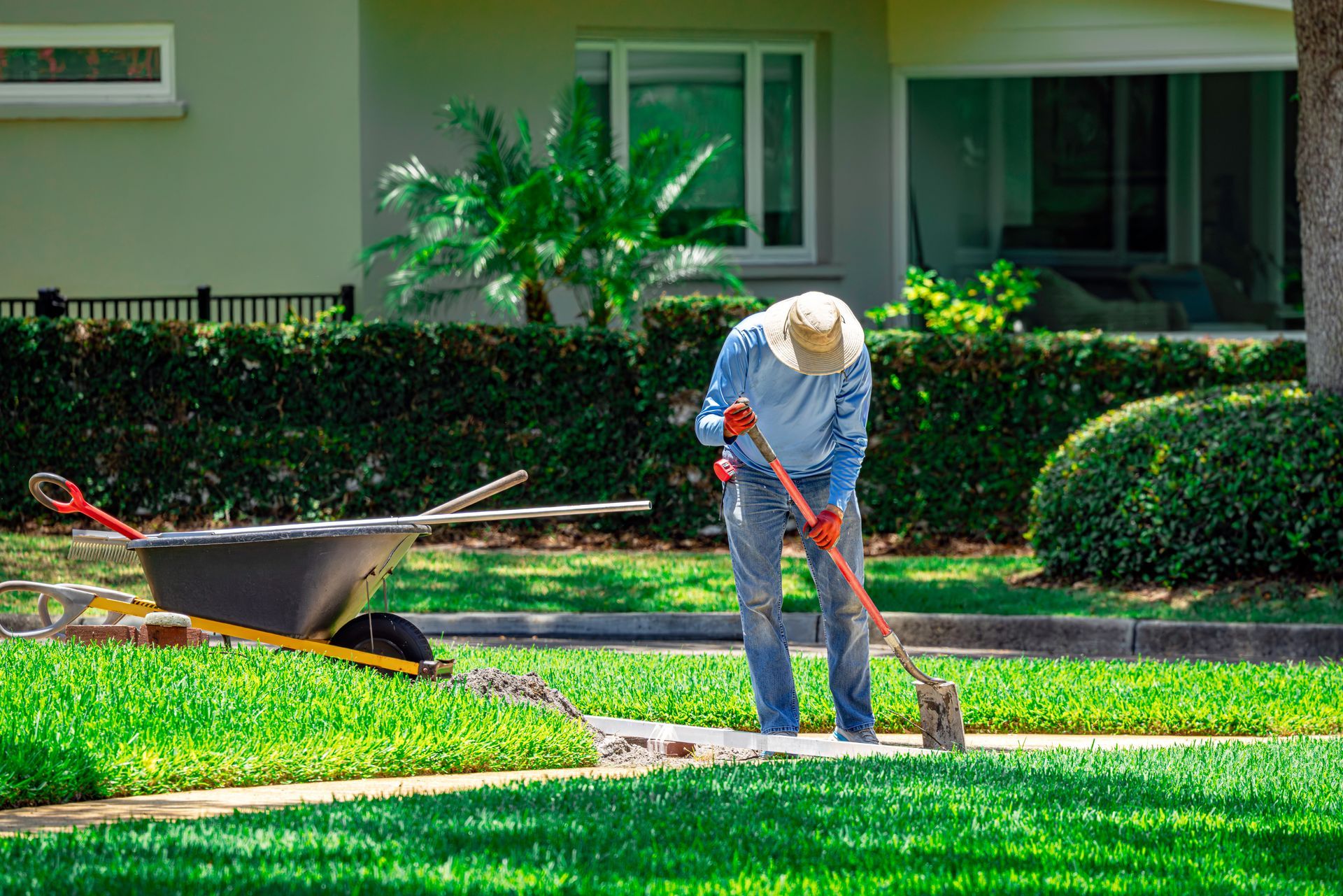 Landscaper in a straw hat working on a lawn edge beside a wheelbarrow in a residential yard