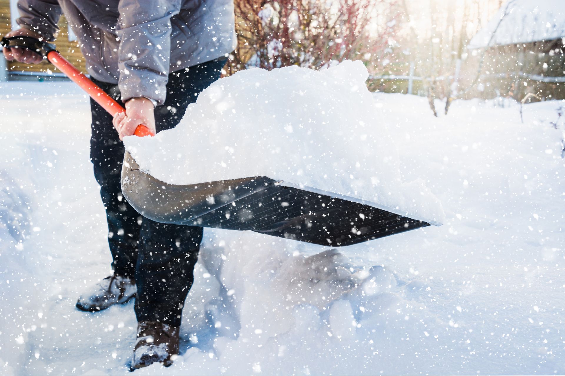 A person in a gray coat and dark pants shoveling deep, white snow during a snowfall.