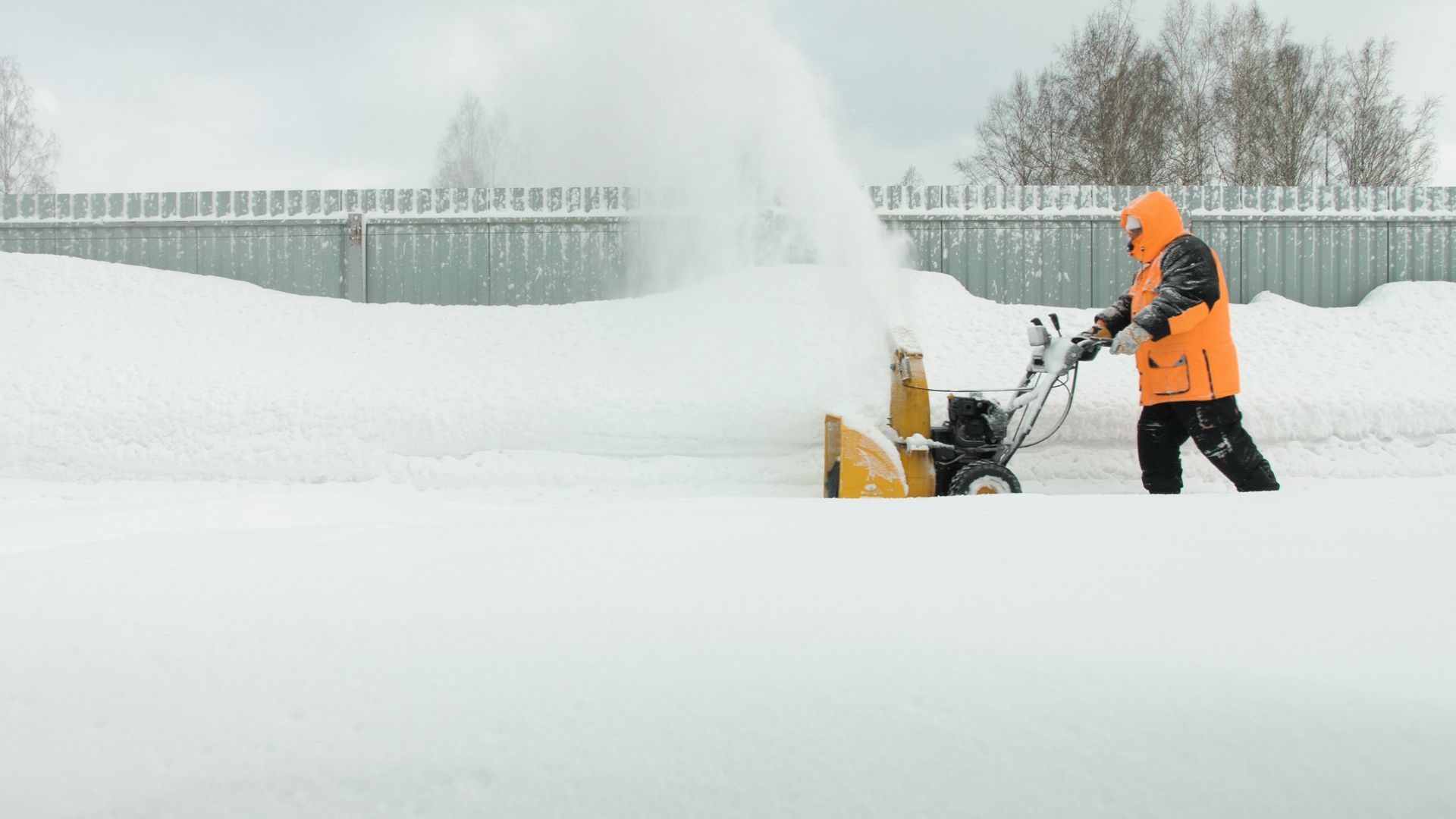 A person in an orange jacket uses a yellow snowblower to clear a path along a fence in a snowy landscape.