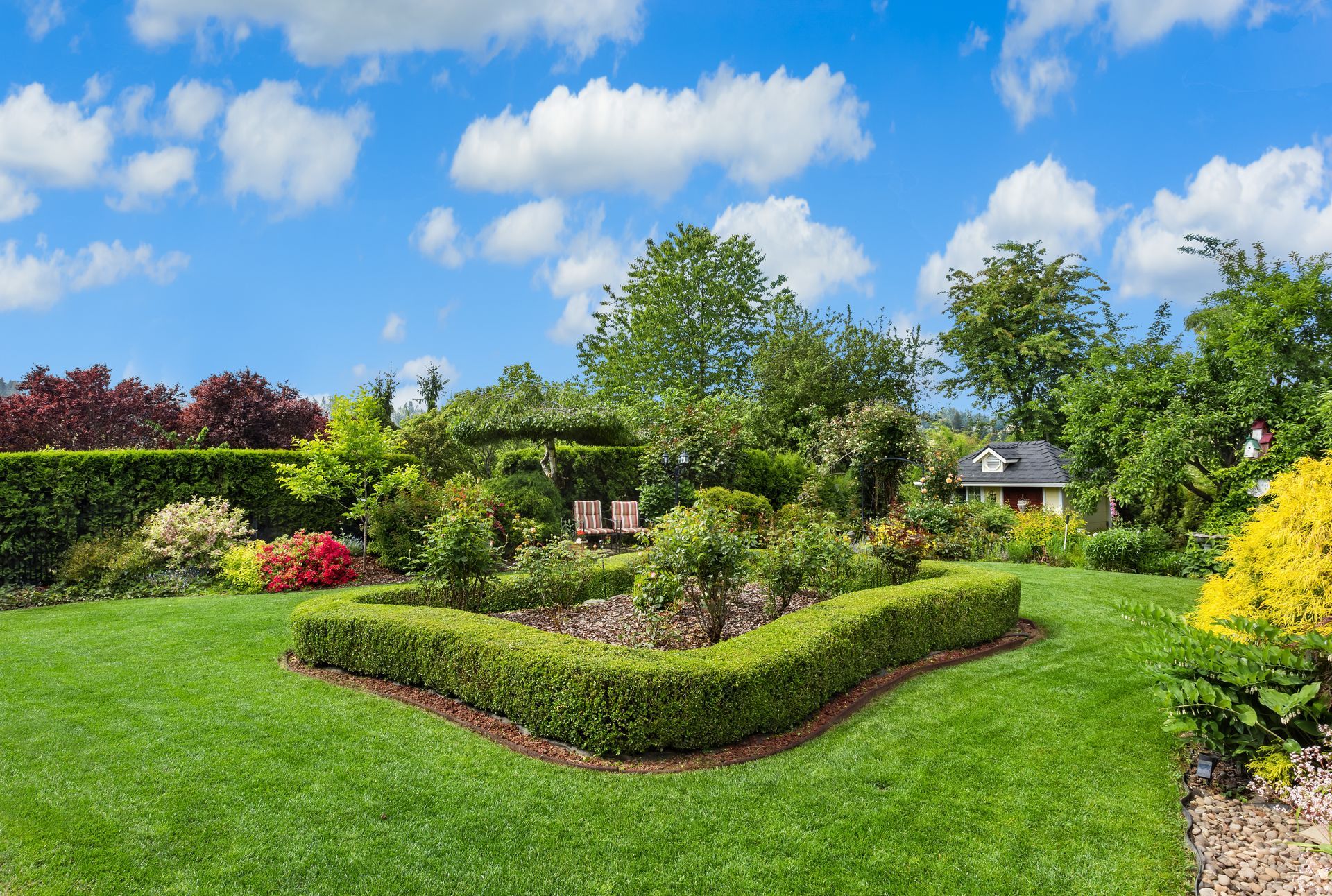 A vibrant green lawn features a manicured boxwood hedge border surrounding a garden bed under a bright, cloudy blue sky.