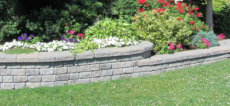 Curved stone garden bed with colorful flowers and green shrubs beside a lawn.
