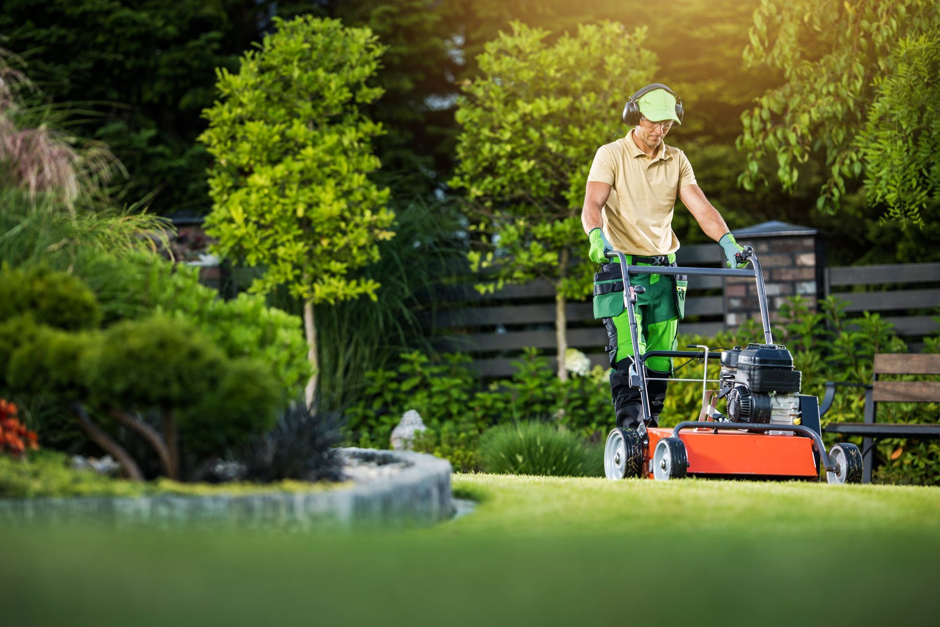 A professional gardener in uniform and protective earmuffs operates a lawn mower in a lush, green landscaped garden.