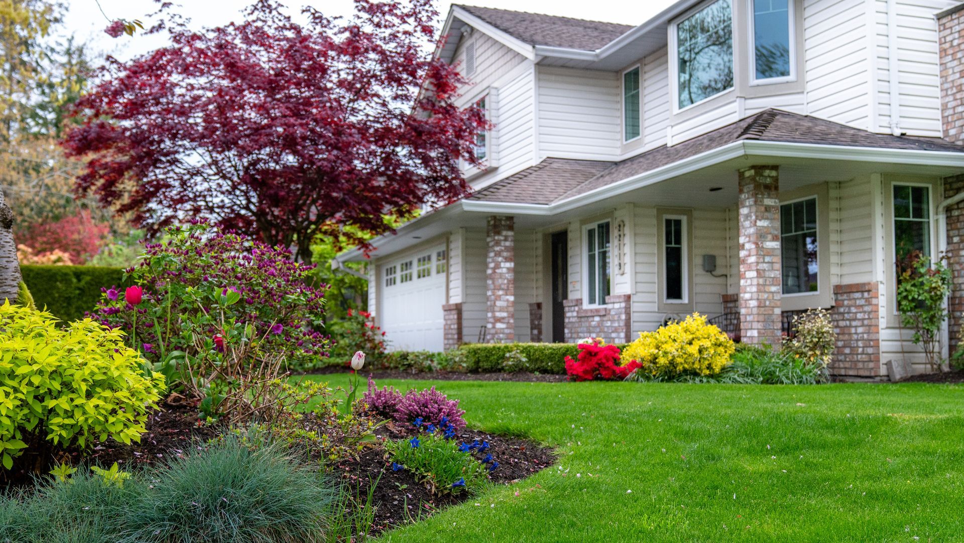 A two-story house with white siding and brick accents, surrounded by a lush green lawn, flower beds, and a red-leaf tree.