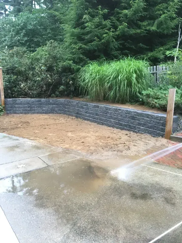 A low stone retaining wall made of stacked grey blocks separates a stone patio from a green lawn and hedge.