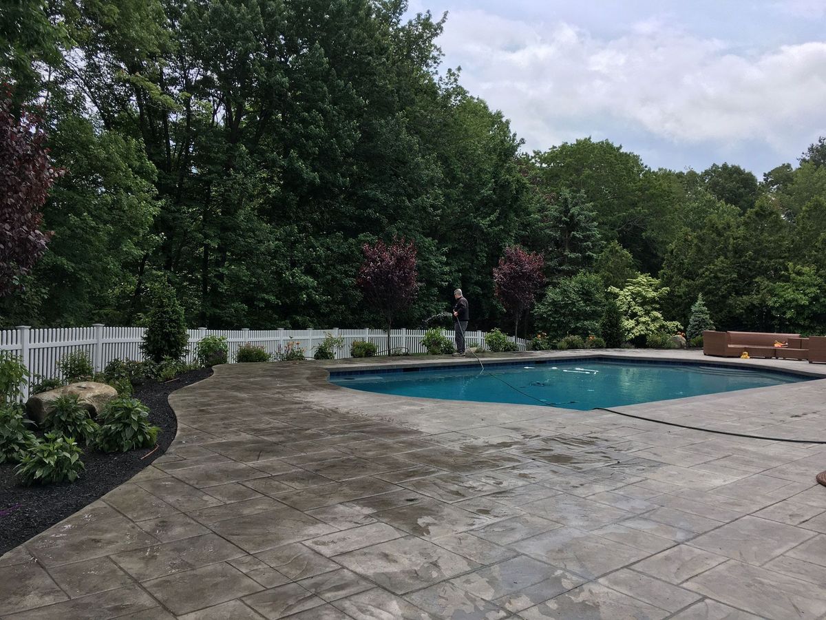 Patio beside a backyard swimming pool with trees, shrubs, and a cloudy sky in the background