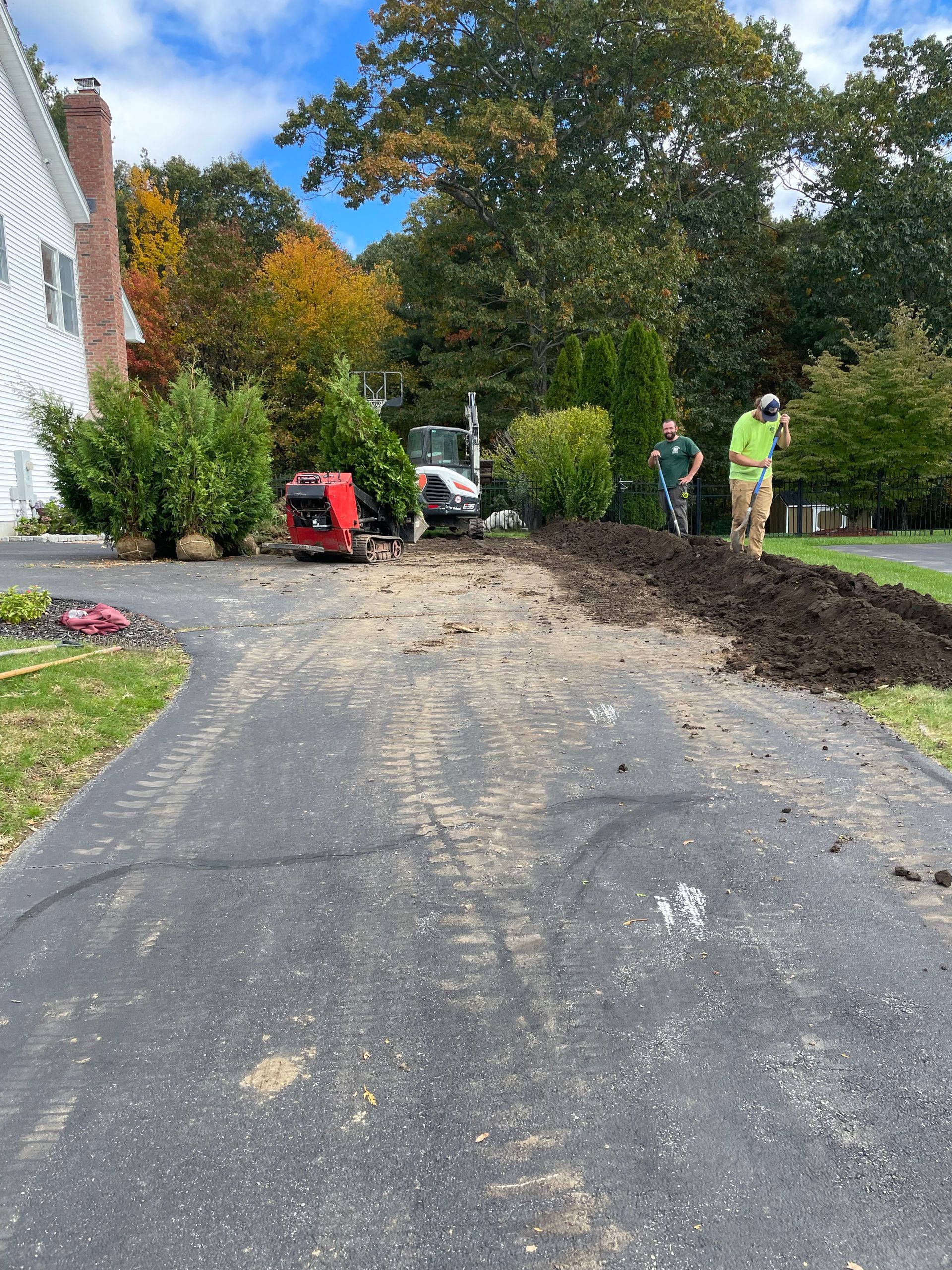 Landscapers use heavy equipment to dig a trench along the edge of a residential asphalt driveway.