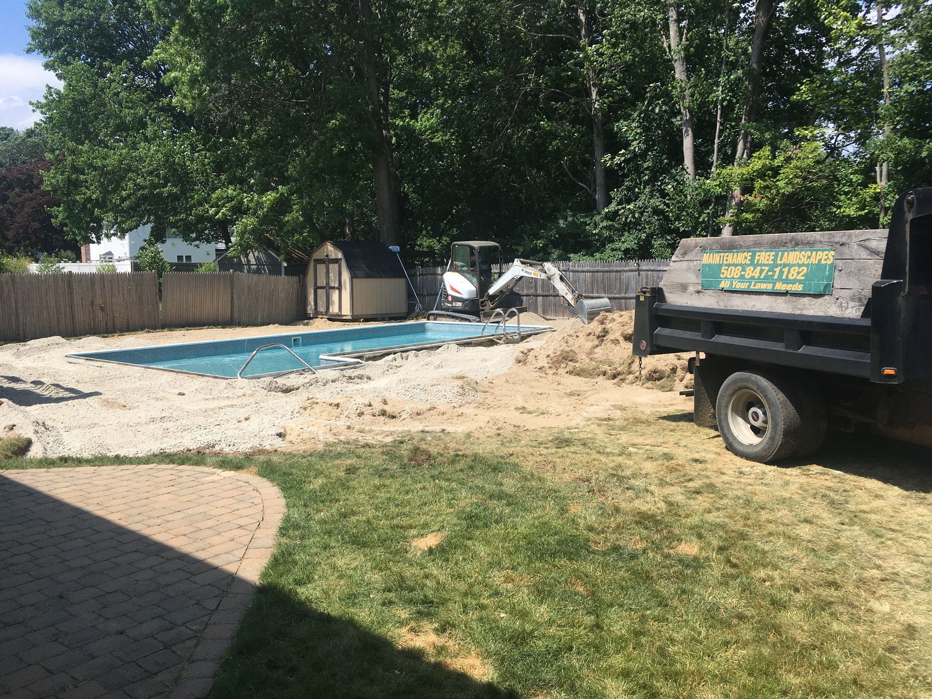 Construction work at a backyard pool with a dump truck parked on grass and a shed near a wooden fence.