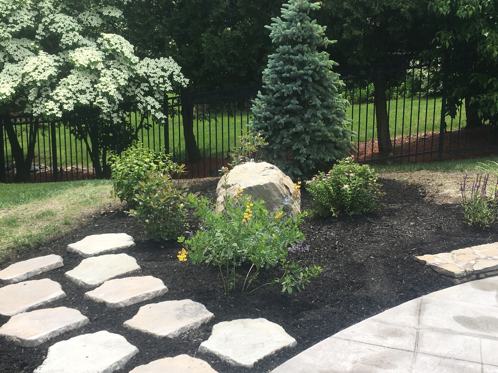 A stone paver path winds through a vibrant green lawn beside a colorful garden bed of red plants and tropical foliage.