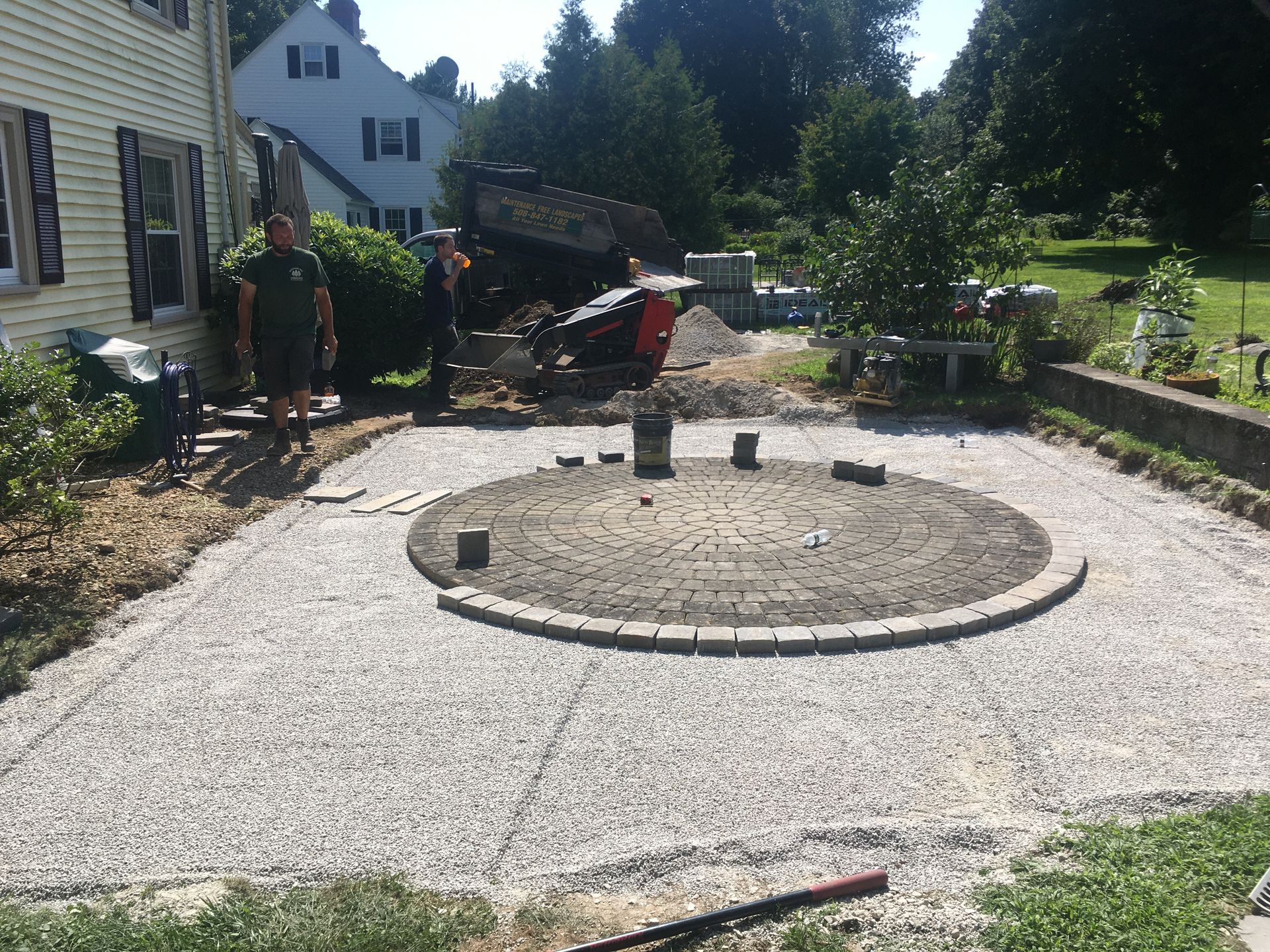 A circular stone fire pit sits on a beige paver patio next to a potted fern with a grassy yard and trees in the background.