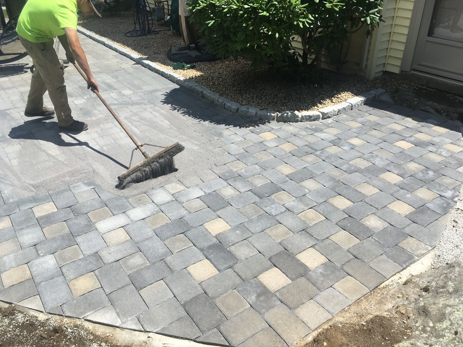 A person in a blue shirt wearing work gloves lays red bricks in a herringbone pattern on a sandy outdoor surface.