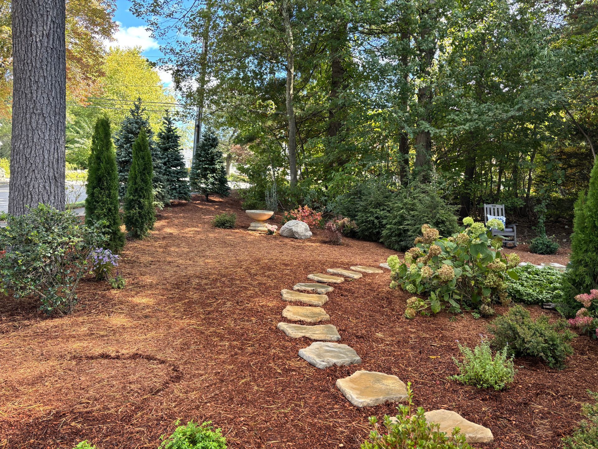 A yellow garden cart filled with dark soil and a shovel sits on a lawn next to a mulched garden bed with hostas.