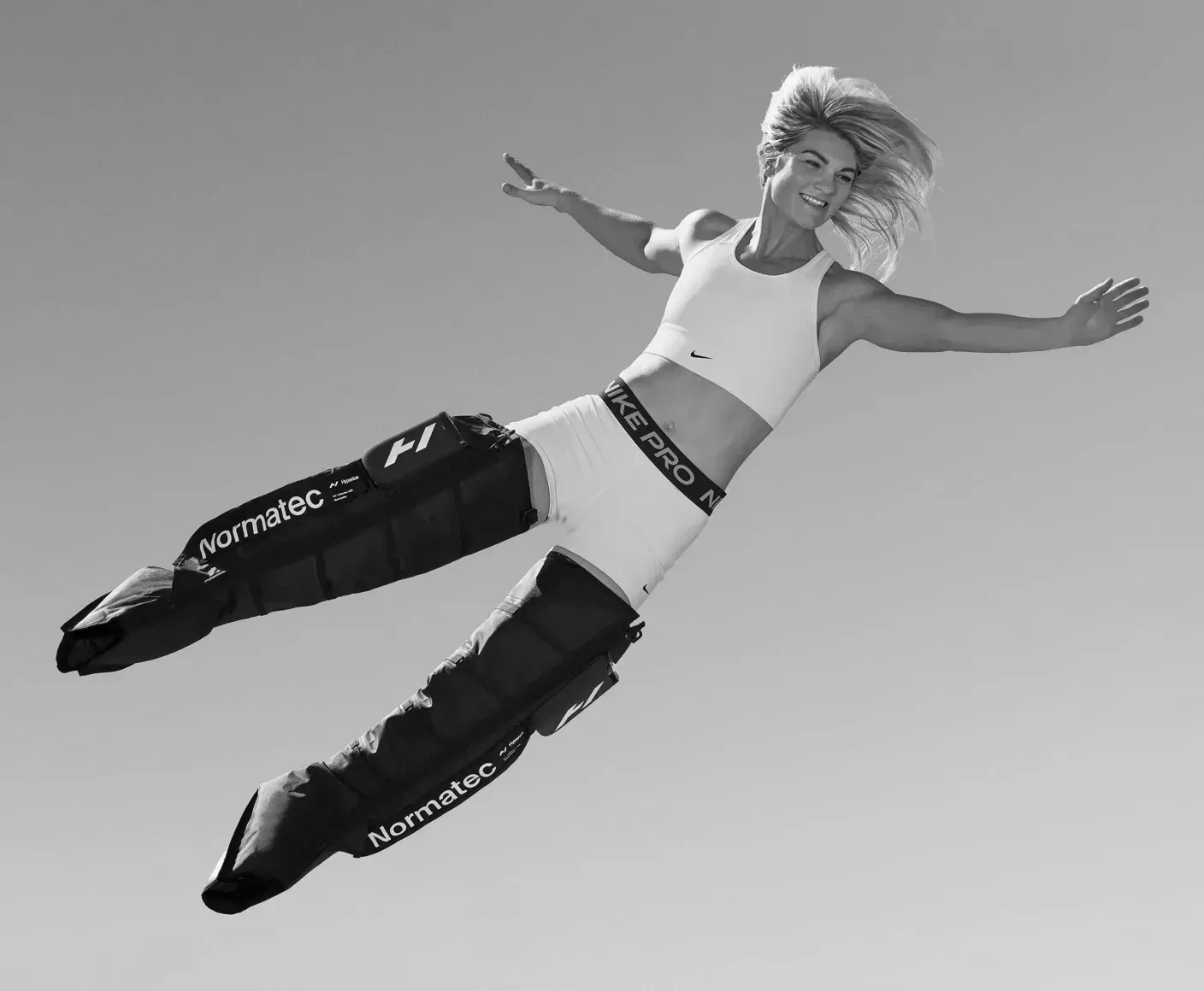 Woman leaping with arms outstretched, wearing sports bra, shorts, and leg compression sleeves against a clear sky.