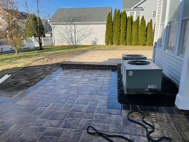 Brick patio beside a house with two AC units and a row of green trees.