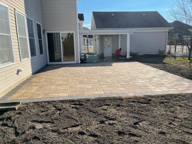 Newly constructed patio paved with light-colored bricks, adjacent to a beige house with large windows.