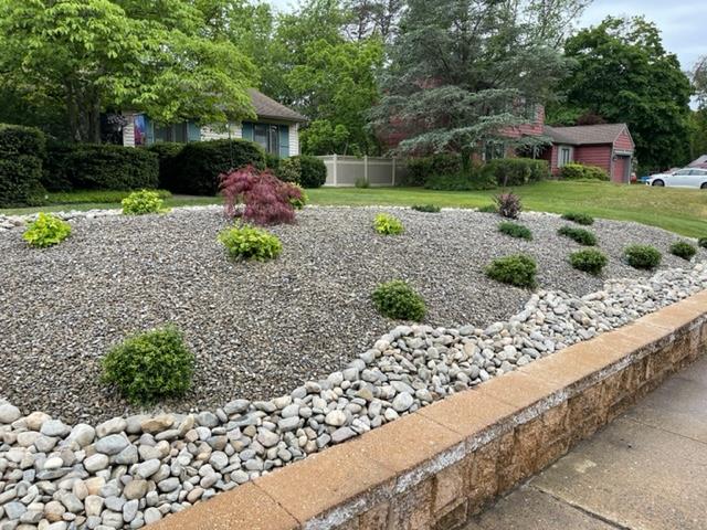 Landscaped garden bed with gray gravel and green shrubs, edged by a brick wall and river rocks.