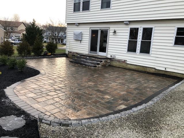 Brick patio with stone border beside a house, steps, and garden bed.
