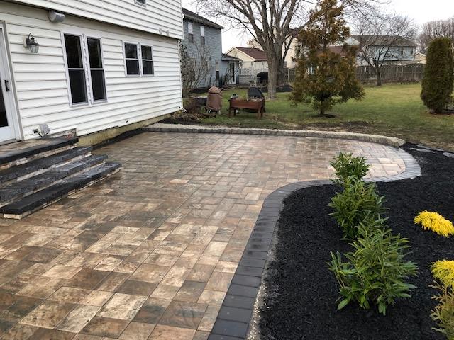 A backyard patio made of paving stones. A dark-bordered garden bed with bushes is on the right. House in background.