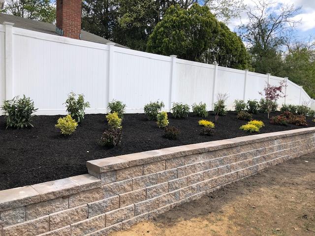 White privacy fence behind a retaining wall with dark mulch and shrubs.