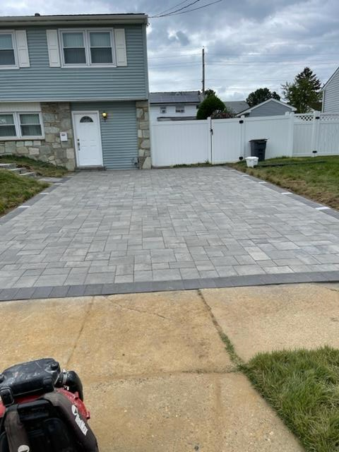 Paver driveway in front of a light blue house. Gray pavers with dark gray border. Compactor in the foreground.