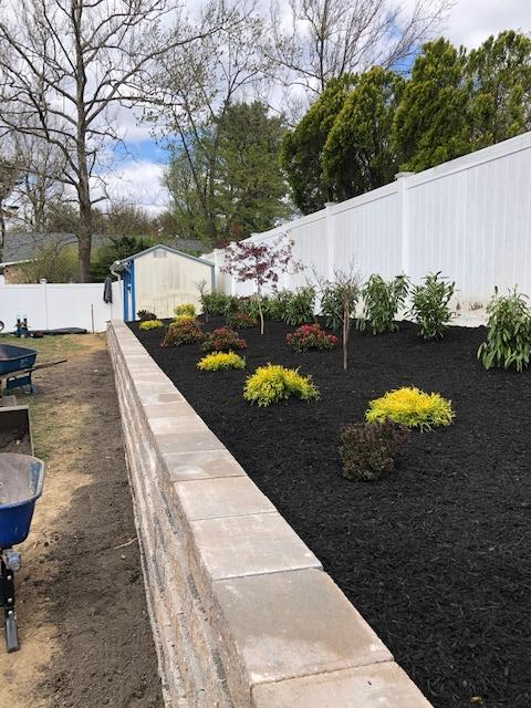Landscaped backyard with retaining wall, mulch, and various plants along a white fence.