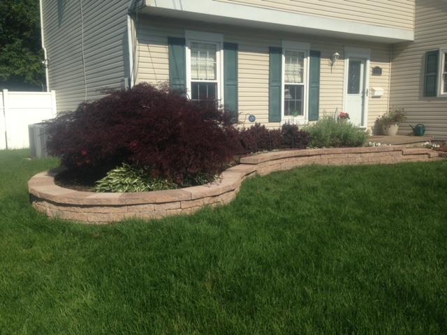 Tan house with green shutters, a curving brick retaining wall, and a dark red bush in front.