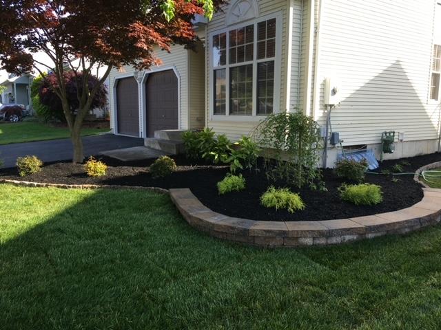 A well-manicured yard features a brick-edged flower bed filled with mulch and green plants, next to a beige house and driveway.