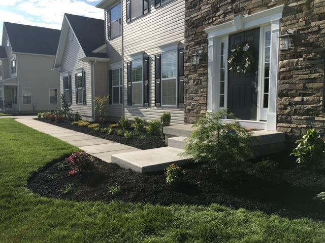 A two-story house with a stone facade entrance, sidewalk, and landscaped garden beds with dark mulch.