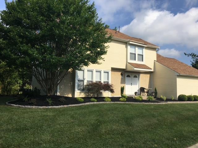 Yellow two-story house with brown roof and lush green lawn, landscaped with black mulch and a large tree.
