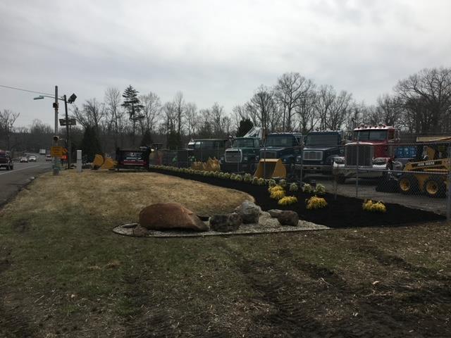 Semi-trucks and construction equipment parked along a roadside, with landscaping in front.