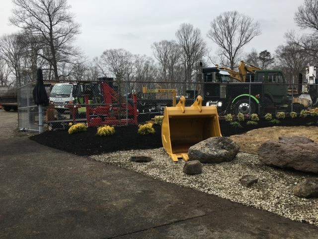 Construction equipment display outdoors with an excavator bucket in foreground. Black mulch and rocks.