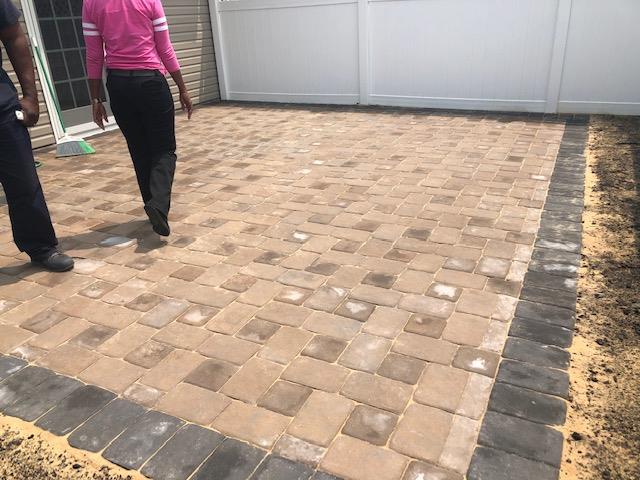 Patio with brick pavers, two people walking across it. Border is a darker brick. White fence in background.