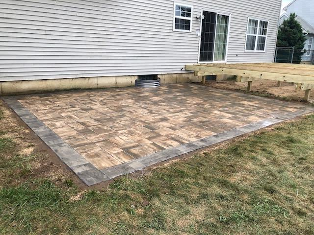 Paver patio with a dark border next to a house with siding and a wooden deck.