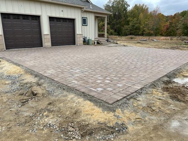 Brick paver driveway in front of a house with brown garage doors; dirt and gravel surround.