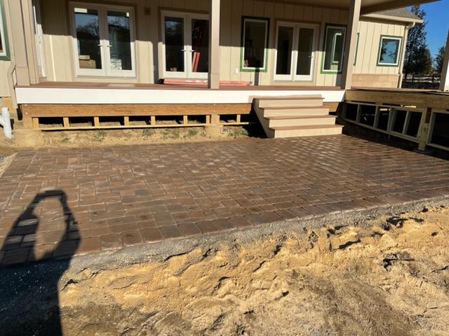 Brick patio under construction next to a house with wooden deck and steps; shadow in foreground.