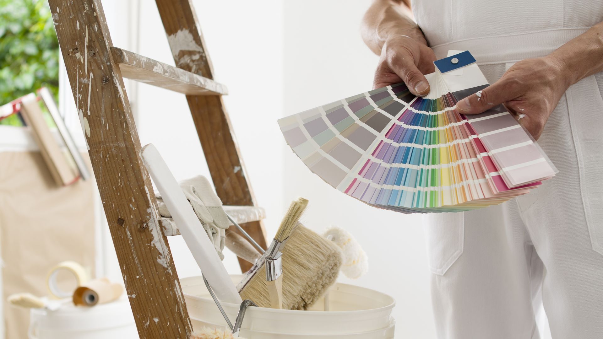 A partial view of a painter holding a colour swatch next to a ladder, paintbrushes, and a bucket.