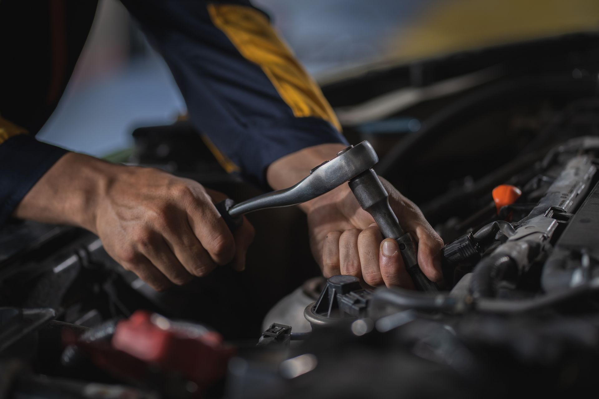 Mechanic's hands working on a car engine with a wrench. Wearing a blue and yellow uniform.