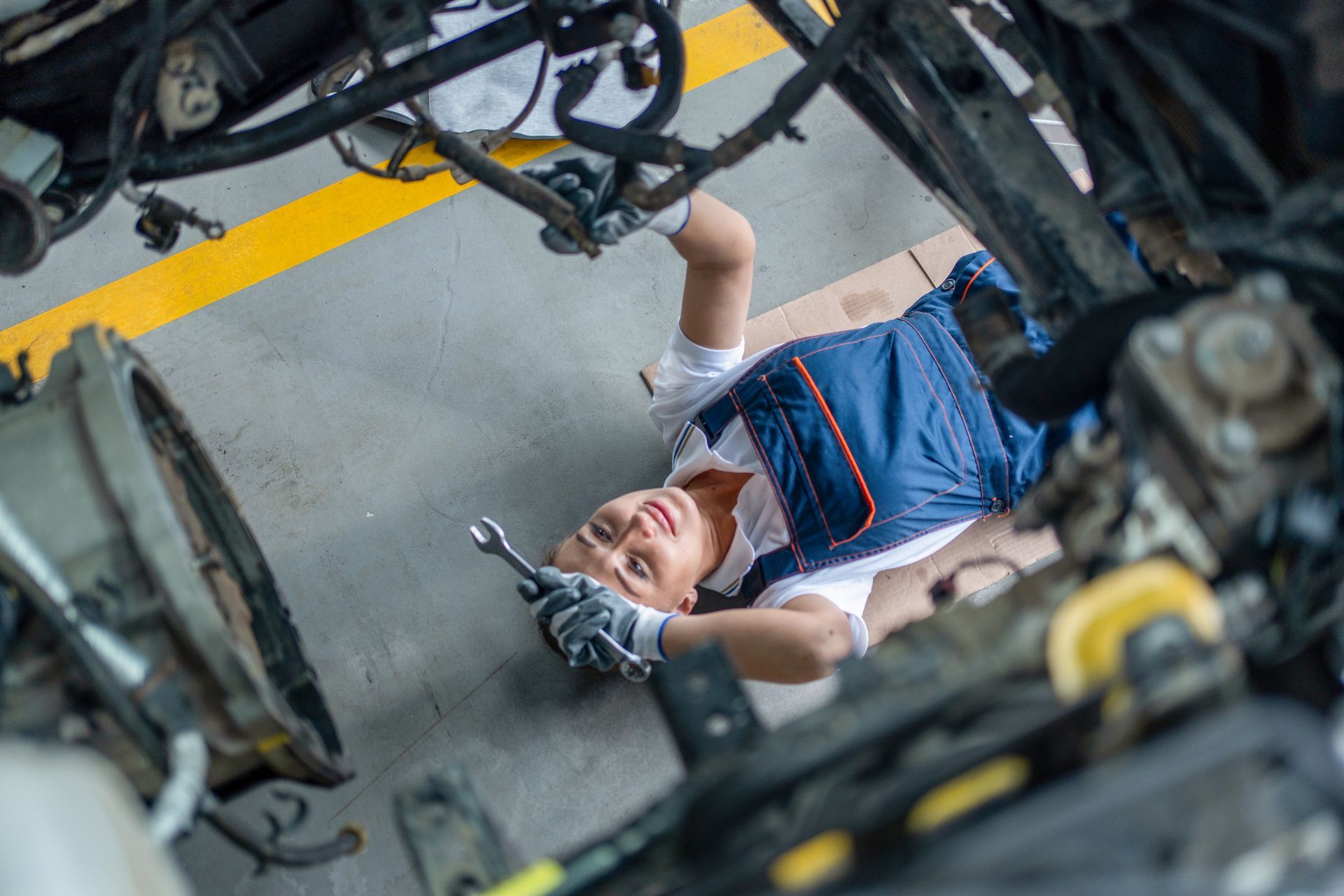 Mechanic in blue coveralls working under a car in a garage. Smiling with a wrench.