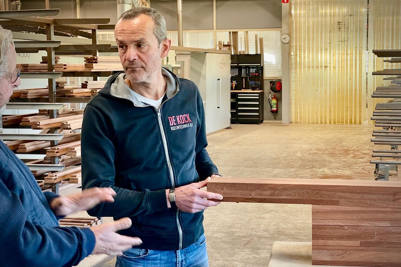 Een man in een zwart jasje praat met een andere man in een houtbewerkingswerkplaats.