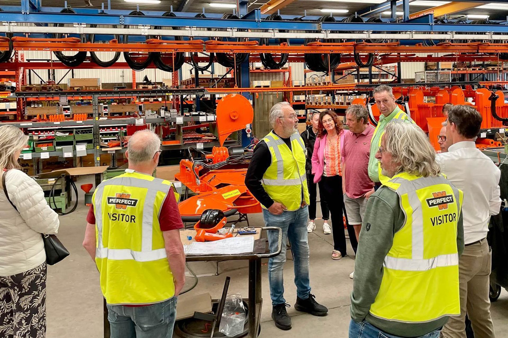 Groepsrondleiding in het magazijn; mensen met veiligheidsvesten staan bij de oranje stellingen met machines en apparatuur.
