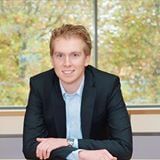 Blonde man in blazer lacht, armen op tafel. Lichtblauw overhemd. Raam met bladeren op de achtergrond.