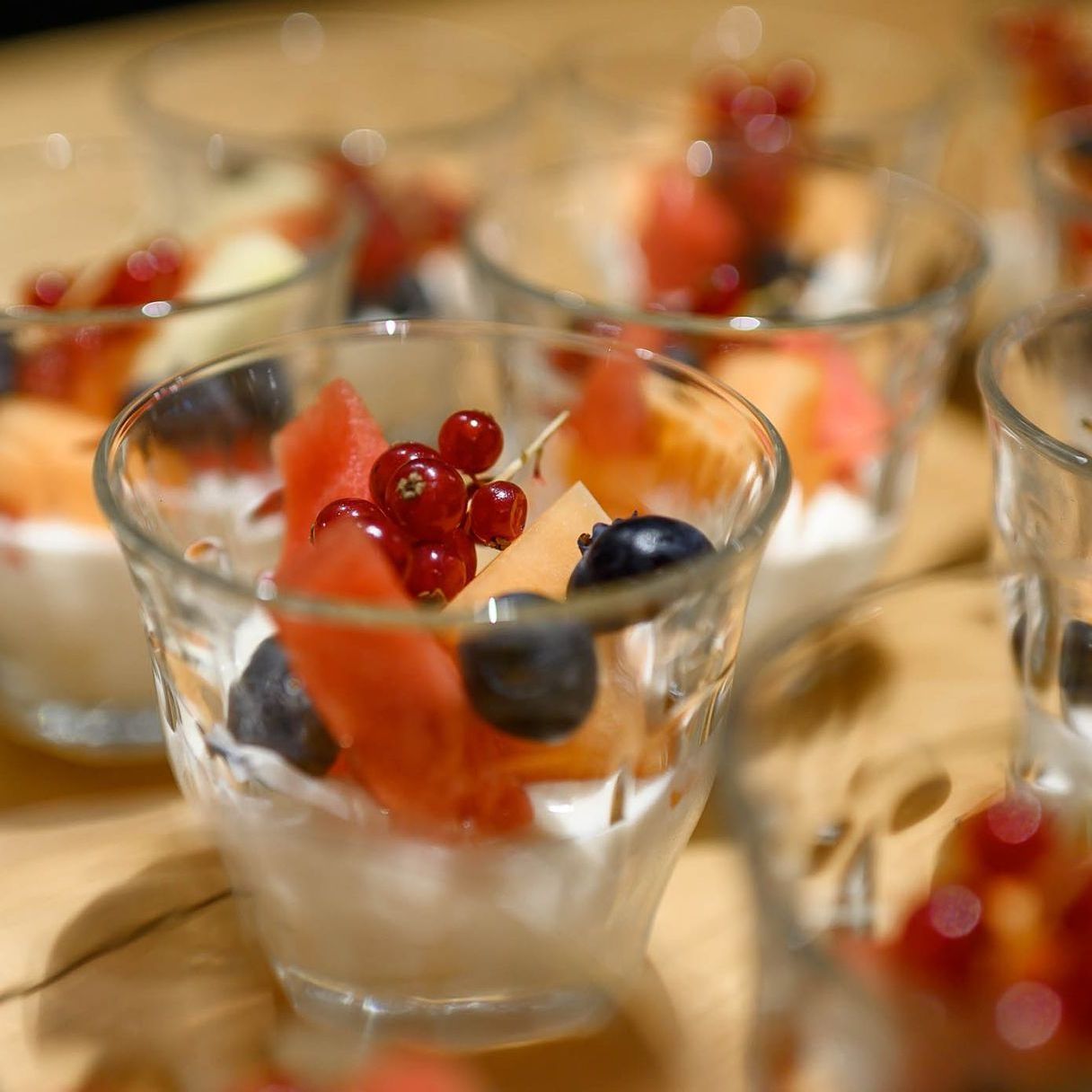 Kleine glazen bakjes gevuld met yoghurt en gegarneerd met fruit: watermeloen, bosbessen en rode bessen.