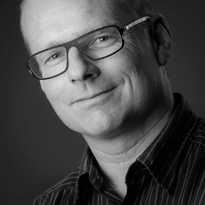 Man met bril en gestreept overhemd lacht, hoofd gekanteld, zwart-wit.