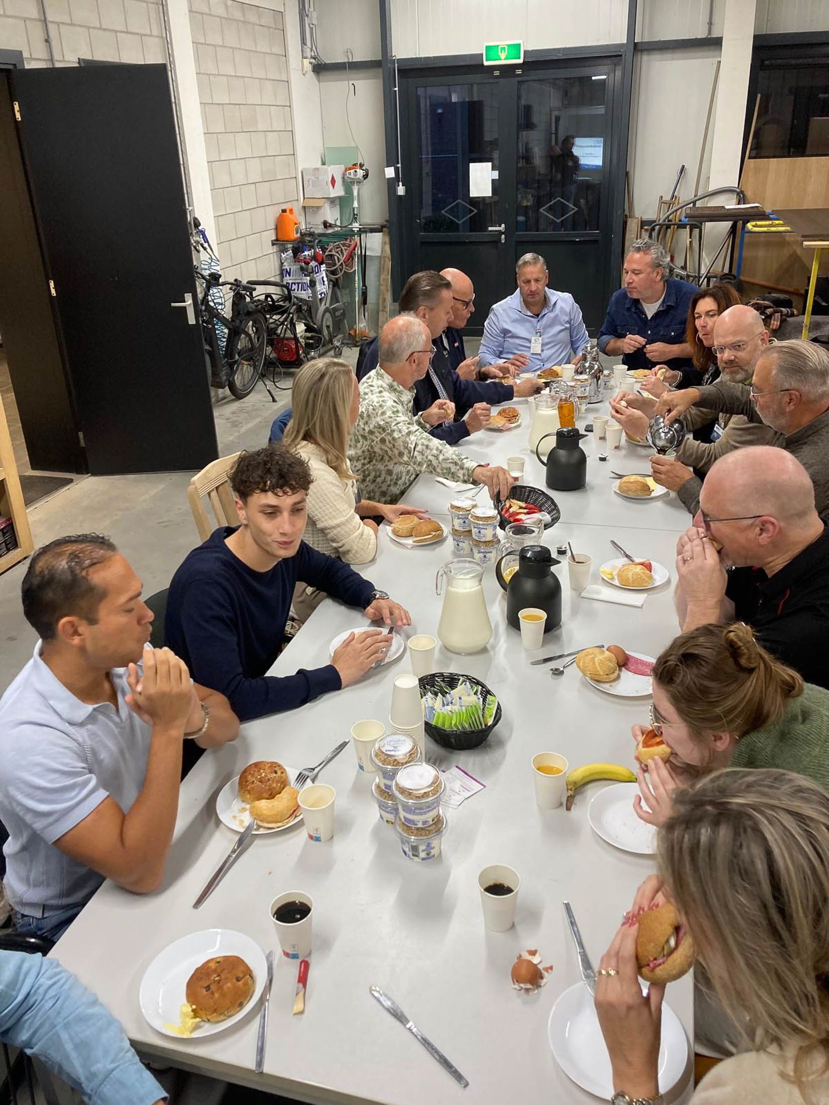 Mensen zitten rond een tafel te ontbijten. Mannen en vrouwen in een industriële setting.