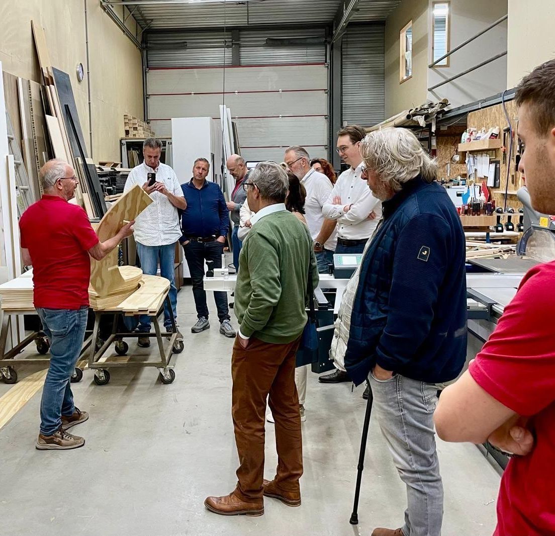 Een groep mensen in een werkplaats. Een man in een rood shirt wijst naar een beeldhouwwerk.