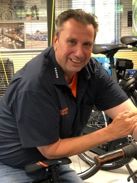 Man in marineblauw overhemd, oranje onderhemd, glimlachend, leunend op een fiets in een winkel.