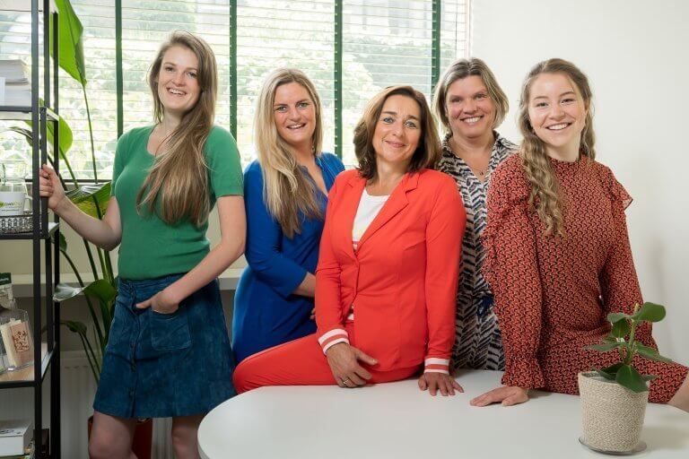 Vijf lachende vrouwen poseren samen binnenshuis, met kleding in verschillende kleuren, bij een raam.